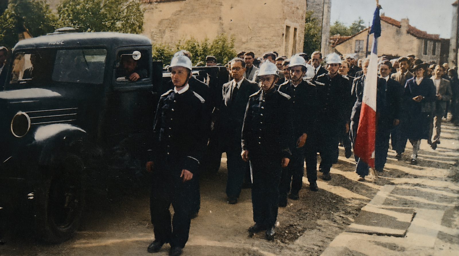 Firemen in polished helmets walk beside the truck carrying the coffins, a French flag behind