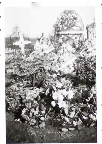 Small black-and-white print: a grave heaped with flowers and wreaths, crosses behind